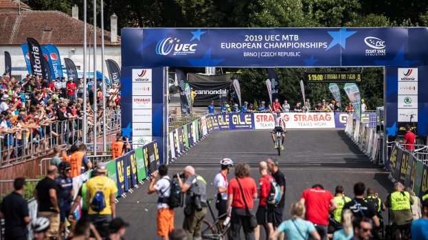 Eine Gruppe von Radfahrern fährt auf einer Straße, die von Zuschauern gesäumt ist, unter einem Bogen hindurch, auf dem "2019 UEC MTB Europameisterschaften" steht, mit Bäumen, Gebäuden und einem klaren blauen Himmel im Hintergrund.