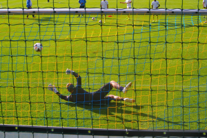 Ein Fussballtorhüter beim Abwehrversuch vor dem Tor, mit einem Netz im Vordergrund und einigen Menschen im Hintergrund, während mehrere Fussbälle auf dem Boden verstreut liegen.