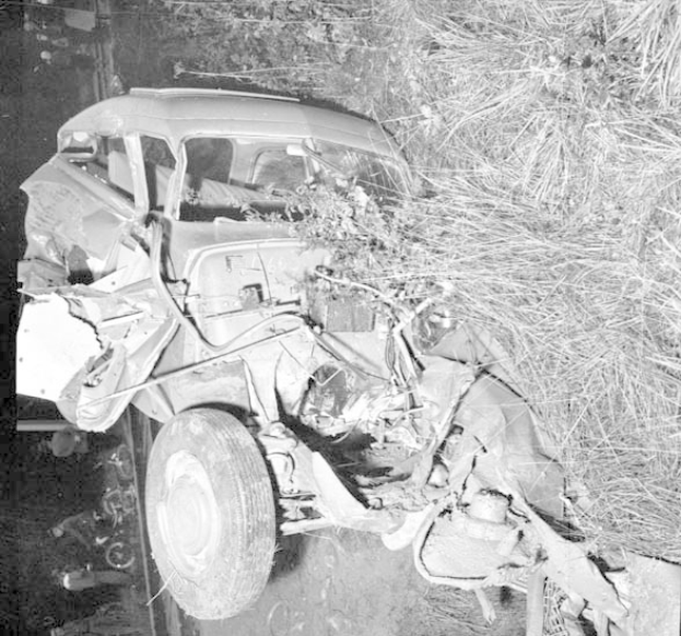 Schwarz-weißes Foto eines schwer beschädigten Autos mit eingedellter Front und zersplitterten Fenstern am Straßenrand, mit einem Baum und verstreuten Gegenständen im Hintergrund.