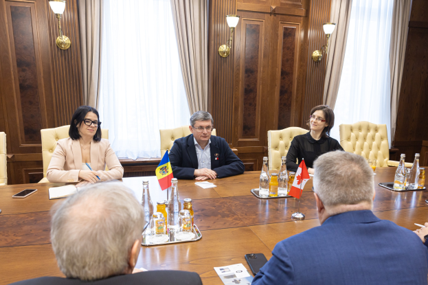 Eine Gruppe von Menschen sitzt um einen hölzernen Tisch in einem formellen Setting, diskutiert mit EU-bezogenen Dokumenten und Gegenständen, darunter Flaggen, Flaschen, Gläser und Mobiltelefone.