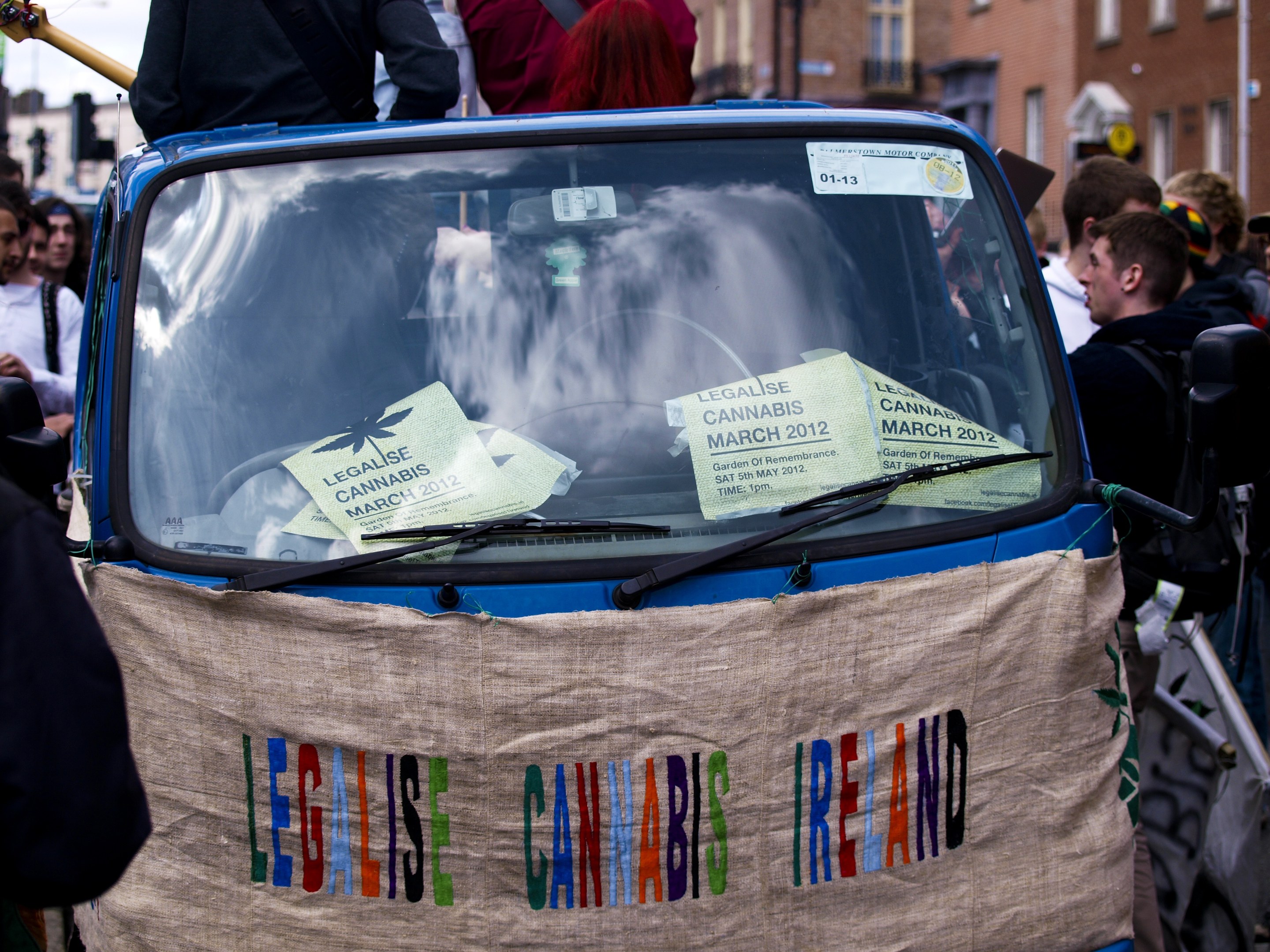 Eine Gruppe von Menschen um ein Auto mit einem "Legalise Cannabis Ireland"-Schild herumstehend, mit Papieren im Auto, umgeben von Gebäuden, Laternenpfählen und einem klaren blauen Himmel im Hintergrund.