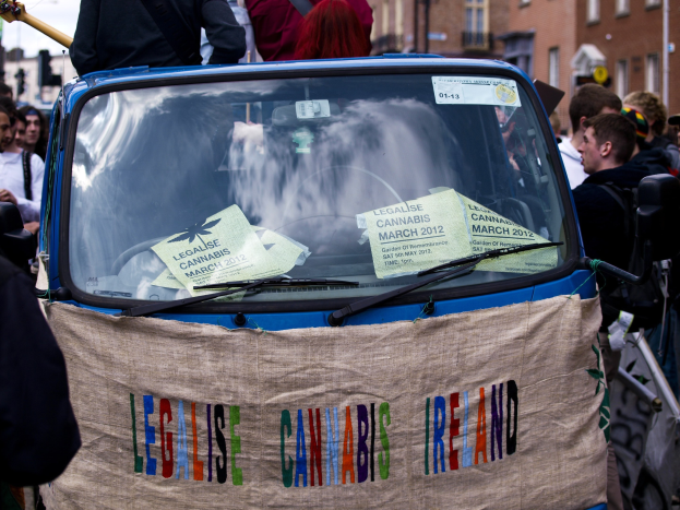 Eine Gruppe von Menschen um ein Auto mit einem "Legalise Cannabis Ireland"-Schild herumstehend, mit Papieren im Auto, umgeben von Gebäuden, Laternenpfählen und einem klaren blauen Himmel im Hintergrund.