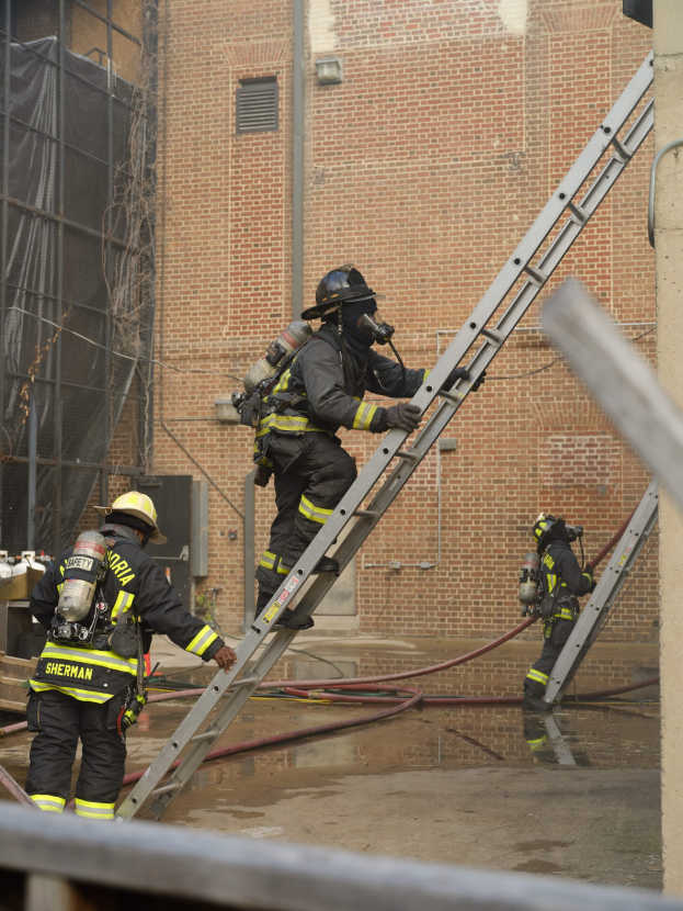 Feuerwehrleute in Helmen und Ausrüstung klettern an einer Leiter vor einem Backsteingebäude mit sichtbaren Rohren und einer Metallstange am Boden, während im Hintergrund ein weiteres Gebäude mit Fenstern und einem Netz zu sehen ist.