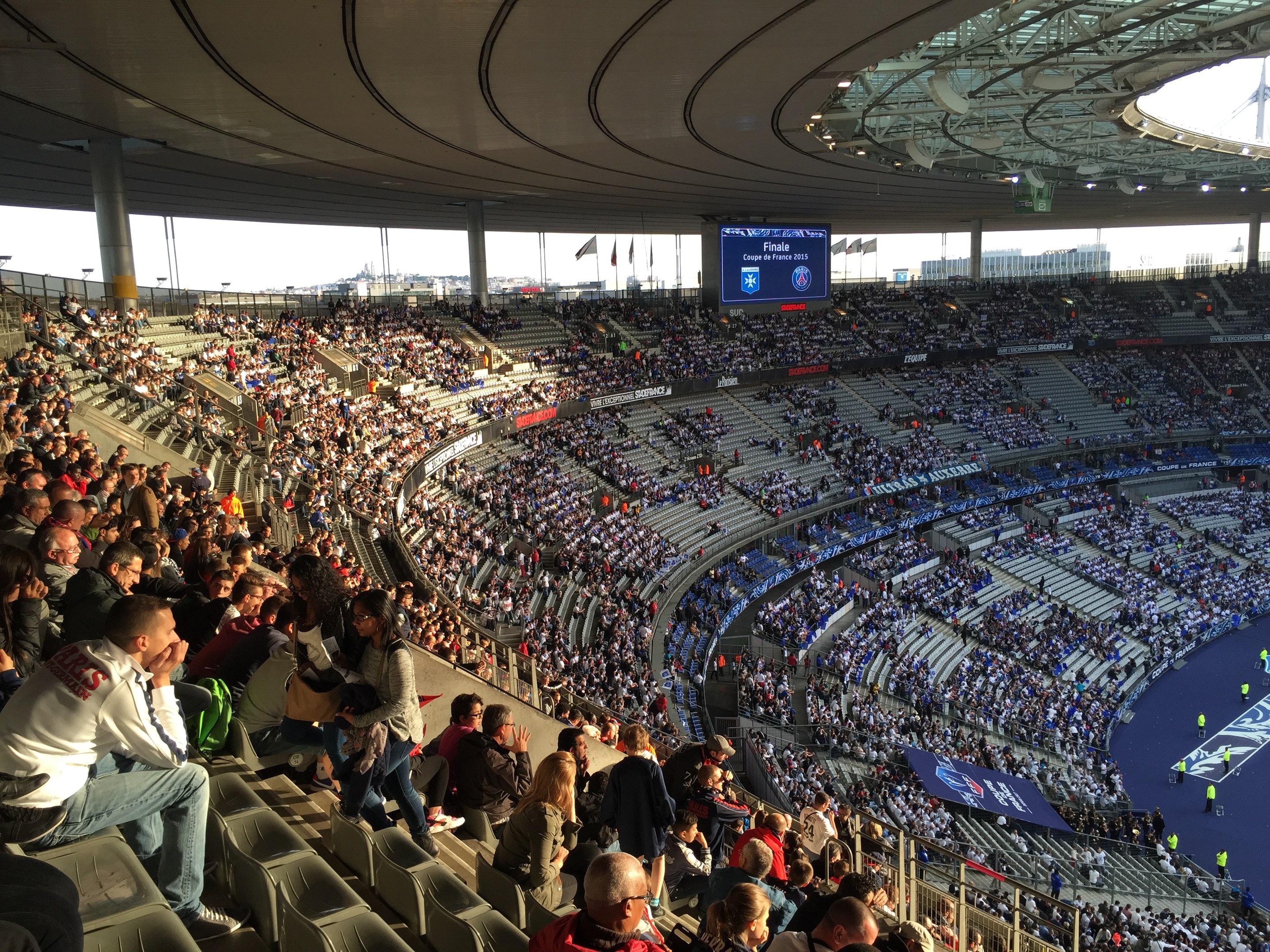 Große Menschenmenge in einem Stadion bei einem Fußballspiel, mit einer Bühne, Fahnen, Stangen und einem Bildschirm im Hintergrund.