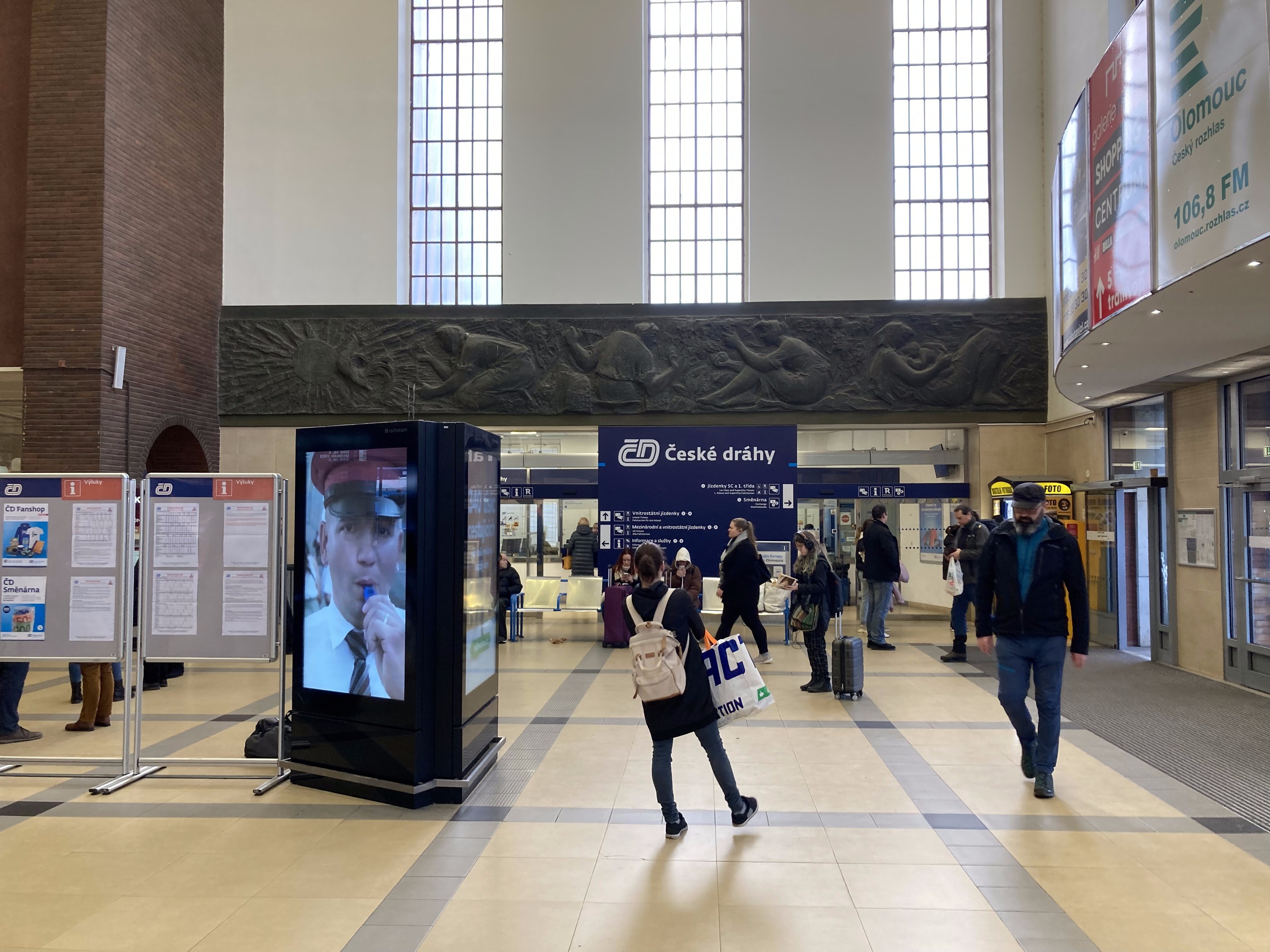 Menschen gehen durch einen Bahnhof mit digitalen Kiosken, Anzeigetafeln und geschnitzten Wänden im Hintergrund.