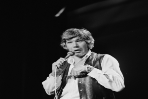 Ein Mann in einem weißen Hemd und schwarzer Weste singt leidenschaftlich in ein Mikrofon, seine Augen sind geschlossen und seine Haare sind im klassischen Rocker-Stil frisiert.