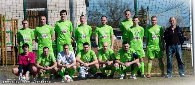 Eine Gruppe von Männern in grünen T-Shirts steht auf einem Fußballfeld in der Nähe eines Torpfostens mit einem Ball auf dem Boden und verschiedenen Hintergrundelementen wie Bäume, Gebäude und Fahrzeuge.