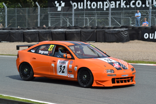 Ein oranges Rennauto mit einem Helmträger am Steuer auf einer Rennstrecke, umgeben von Reifen, Absperrungen, Zuschauern, Bäumen und einem Banner im Hintergrund.