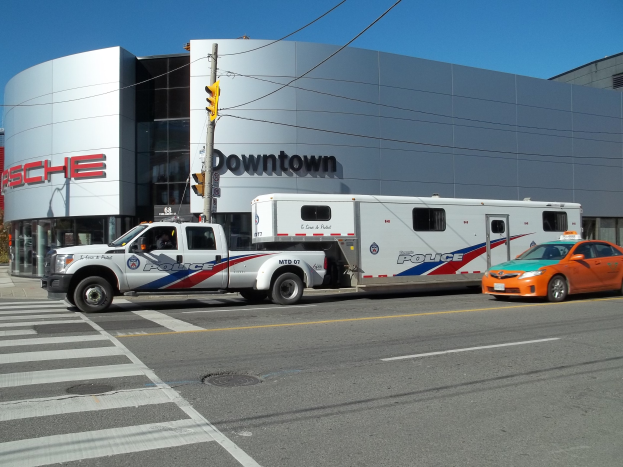 Polizeifahrzeug vor einem Gebäude mit Text geparkt, mit einer Ampel und Kabeln im Vordergrund, Bäume und einen klaren blauen Himmel im Hintergrund.