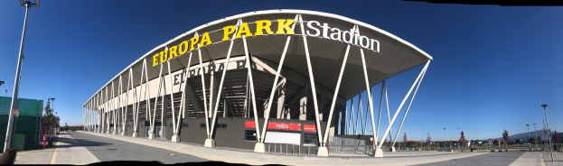 Großes Stadiongebäude mit Text darauf, umgeben von Pfählen, Lampen, Bäumen und einem Zaun, mit Himmel im Hintergrund.