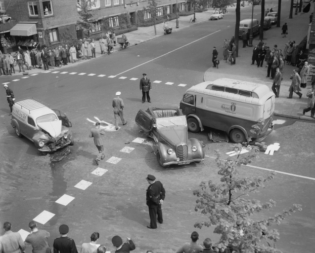 Schwarz-weiß-Szene eines Autounfalls an einer Straßenkreuzung mit einem Lieferwagen, anderen Fahrzeugen und einer Gruppe von Schaulustigen; Gebäude, Bäume, Pfähle und Texttafeln im Hintergrund.