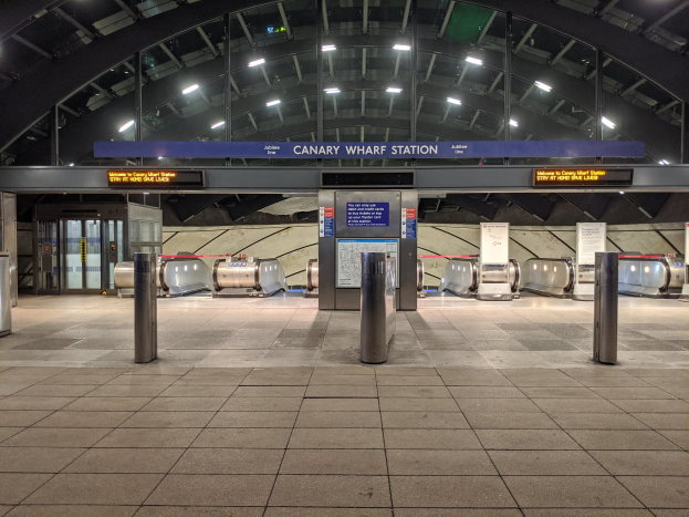Ein U-Bahnhof-Innenraum mit Metalldetektoren auf dem Boden, Pollern, Informationsschildern mit der Aufschrift "Canary Wharf Station", Türen, Wänden und Deckenleuchten.