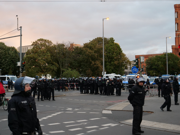 Eine Gruppe von Polizisten in Helmen steht auf einer Straße umgeben von Fahrzeugen, Verkehrsampeln, Laternen, Bäumen, Gebäuden und einem bewölkten Himmel.