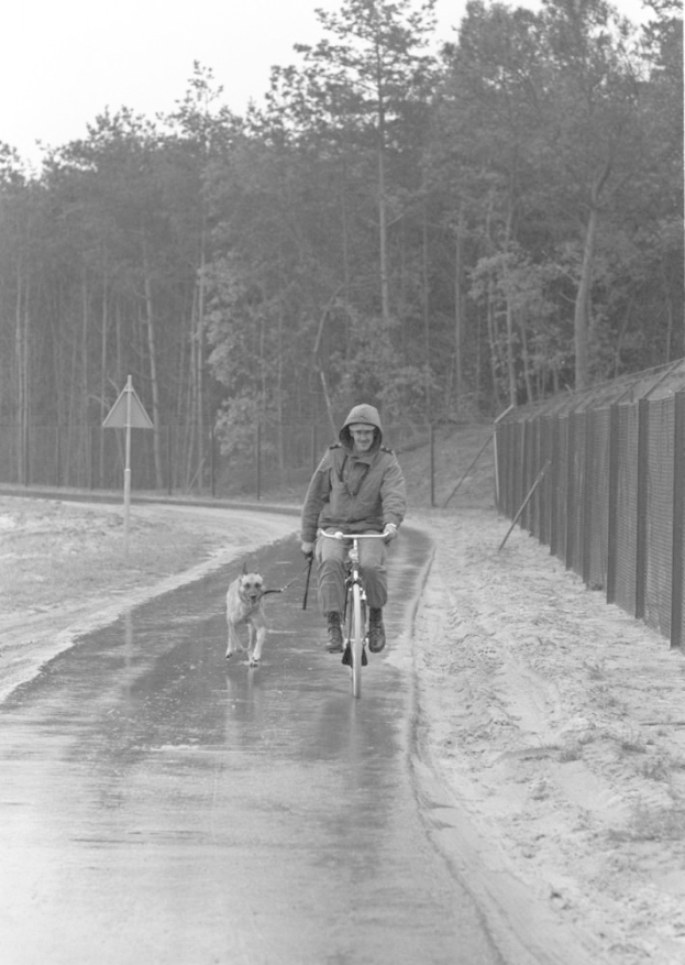 Ein Mann in einer Jacke und einer Mücke fährt ein Fahrrad auf einer nassen Straße mit einem Hund an der Leine vorbei an einem Zaun auf der rechten Seite, mit Bäumen und einem Schild im Hintergrund unter einem sichtbaren Himmel.