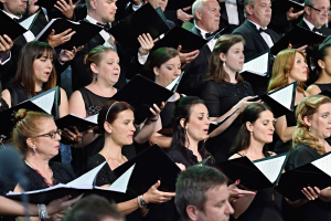 Eine Gruppe von Menschen steht vor einem Chor und hält jeweils ein Buch in der Hand, scheinbar im Einklang singend.