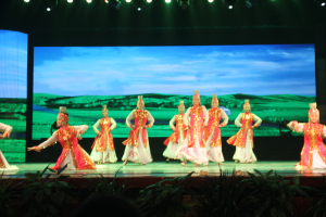 Eine Gruppe von Menschen in bunten Kostümen performs auf einer Bühne, die von Scheinwerfern beleuchtet wird und von Pflanzen umgeben ist, mit einem Bildschirm im Hintergrund.