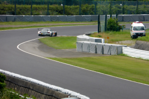 Ein Rennauto fährt auf einer Rennstrecke mit einem Polizeiwagen im Hintergrund, umgeben von Gras, Pflanzen, Bäumen und Absperrungen.