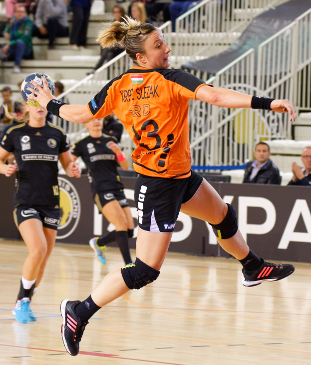 Eine Frau in einem orangen T-Shirt und schwarzen Shorts hält einen Handball, mit Zuschauern auf Treppen und Geländern im Hintergrund während der "Krp Slezak - Handball-Weltmeisterschaft 2019."