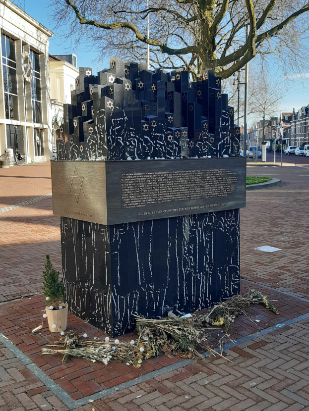 Denkmal in einem Stadtplatz, das Holocaust-Opfern gewidmet ist, mit einem Baum davor, einem Topf mit einer Pflanze auf dem Boden, verstreuten trockenen Blättern und im Hintergrund Gebäude, Fahrzeuge und Bäume.