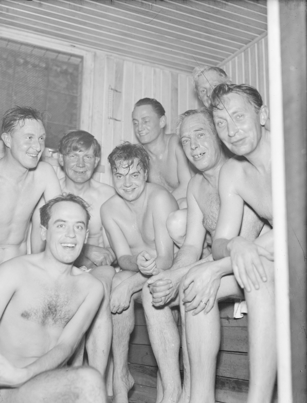 Eine Gruppe von Männern sitzt auf einer hölzernen Bank in einer Sauna, mit einer Wand und einem Fenster im Hintergrund, dargestellt in Schwarz-Weiß.