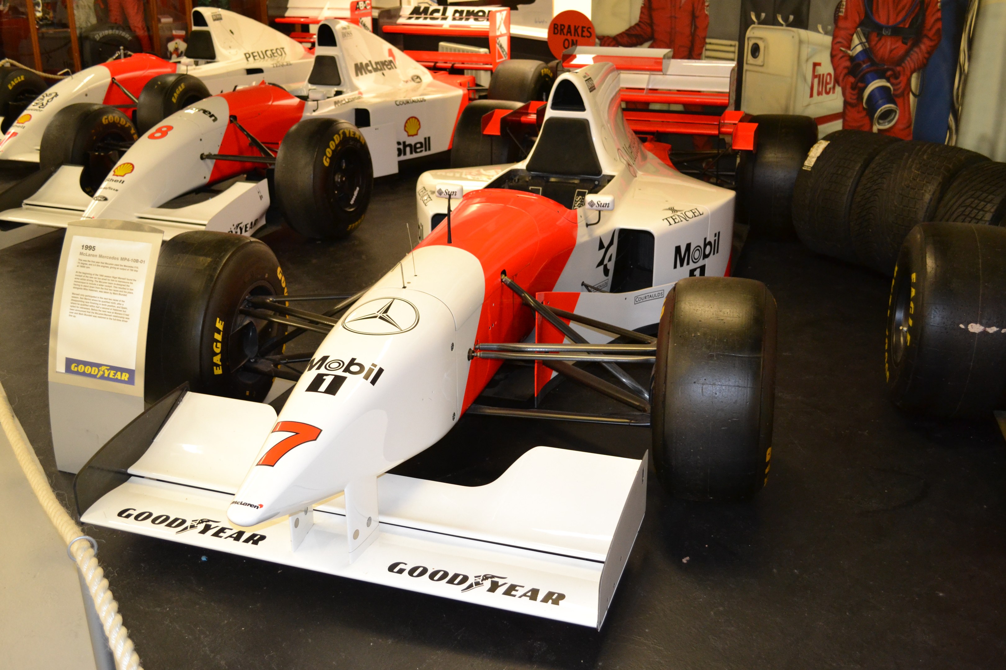 Rennwagen in einem Museum ausgestellt, mit Besuchern, die sie hinter einer Seilabsperrung betrachten, mit Reifen und Rädern und einer Texttafel im Hintergrund.