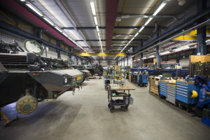 Großer Lagerraum mit Maschinen, Fahrzeugen auf der linken Seite, Transportwagen mit Gegenständen und Holzboxen auf der rechten Seite und Deckenbeleuchtung.