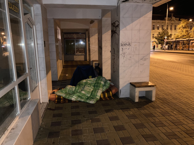 Eine obdachlose Person, die auf einem Bürgersteig in der Nacht schläft, mit einer Decke und einem Hocker in der Nähe, vor einem Gebäude mit einer Schautafel, einem Straßenschild, Bäumen, Fahrzeugen, Passanten und einem bewölkten Himmel.