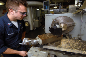 Ein Monteur mit Schutzbrille bedient eine Maschine in einer Fabrikumgebung, mit Industrieausrüstung, Rohren, Beleuchtung und einem Bildschirm im Hintergrund.