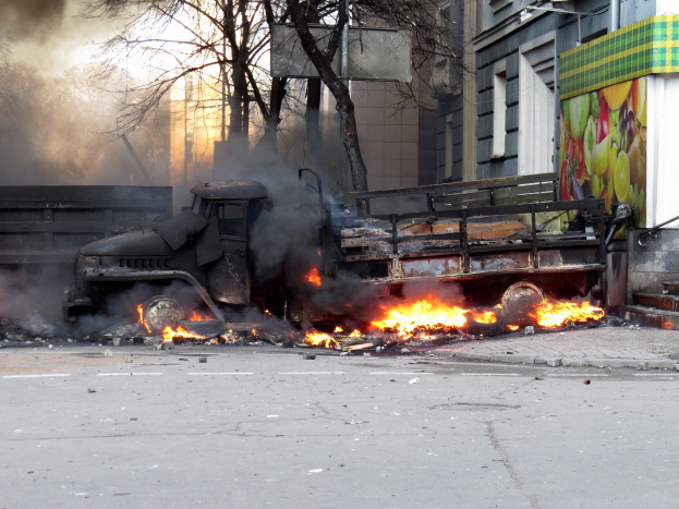 Ein großer Lastwagen brennt auf der Seite einer Straße mit dicken schwarzen Rauchwolken, umgeben von Bäumen, Gebäuden und einem klaren Himmel im Hintergrund.
