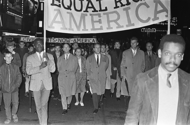 Schwarz-weißes Foto einer Gruppe von Menschen, die eine Straße entlang marschieren und ein Banner mit der Aufschrift "Gleiche Rechte Amerika" tragen, mit Gebäuden und Straßenlaternen an der Straße.