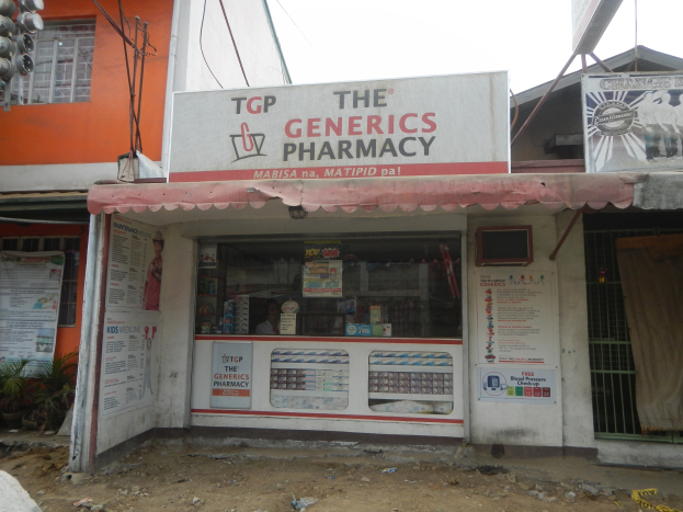 Außenansicht der "The Generics Pharmacy"-Filiale mit umliegenden städtischen Elementen unter einem klaren blauen Himmel.