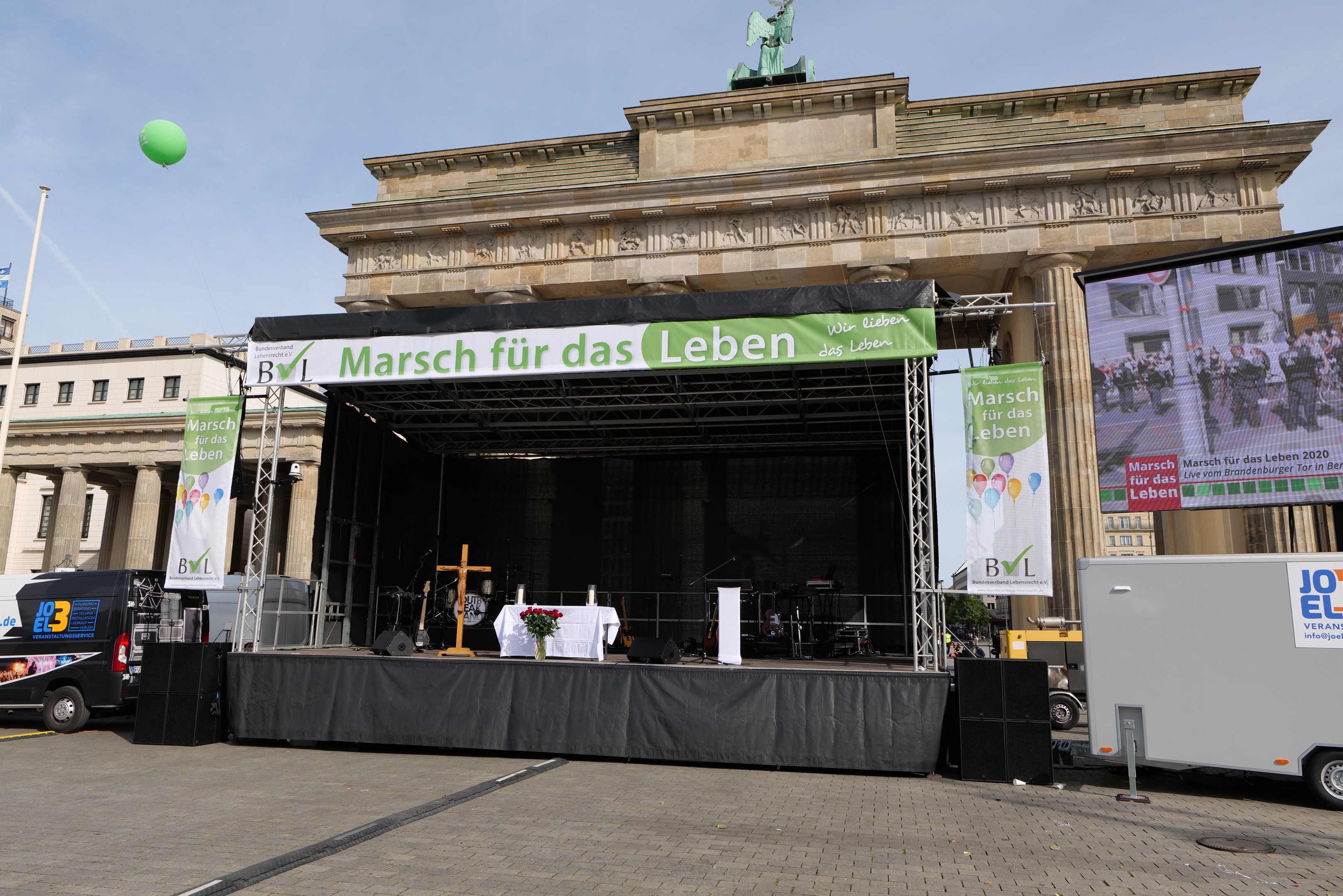 Bühne vor dem Brandenburger Tor in Berlin mit einem Tisch, einer Vase mit Blumen, einem Kreuzsymbol und Bannern sowie Lautsprechern und anderem Equipment; Fahrzeuge auf der Straße und Gebäude mit Fenstern im Hintergrund.