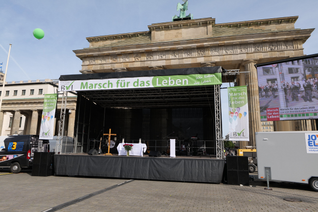 Bühne vor dem Brandenburger Tor in Berlin mit einem Tisch, einer Vase mit Blumen, einem Kreuzsymbol und Bannern sowie Lautsprechern und anderem Equipment; Fahrzeuge auf der Straße und Gebäude mit Fenstern im Hintergrund.