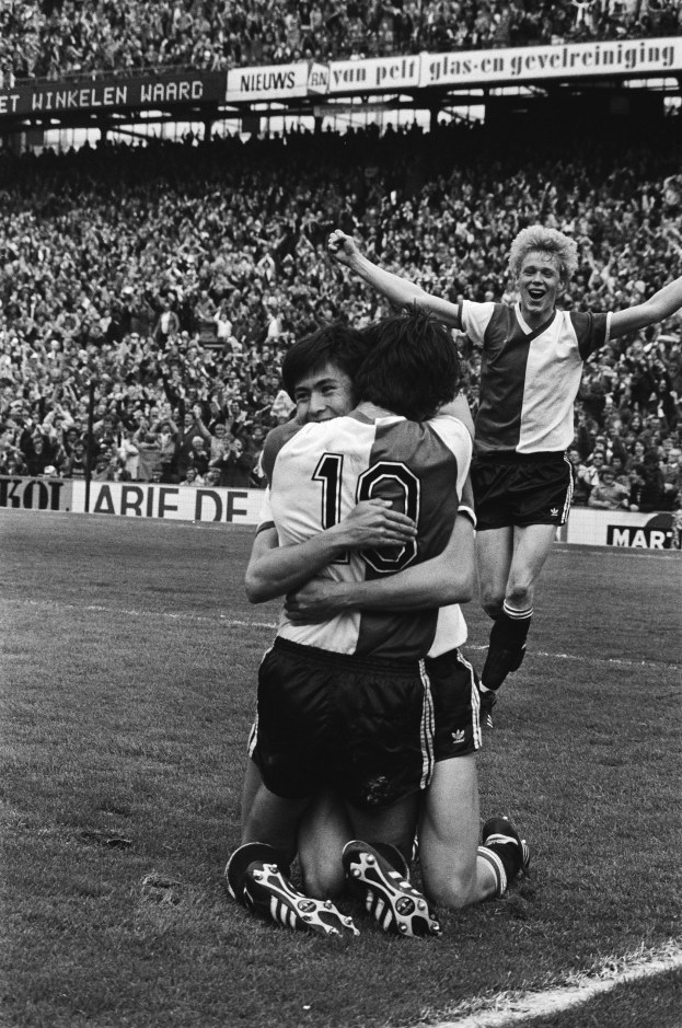 Drei Fußballspieler feiern ein Tor auf dem Boden, indem sie sich umarmen, vor einem jubelnden Publikum und schwenkenden Fahnen im Hintergrund, dargestellt in Schwarz-Weiß.