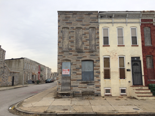 Ein altes, baufälliges Gebäude an einer Straßenecke mit kaputten Fenstern, bröckelnden Wänden, vorbeifahrenden Fahrzeugen, Mülltonnen auf dem Gehweg und einem klaren blauen Himmel.