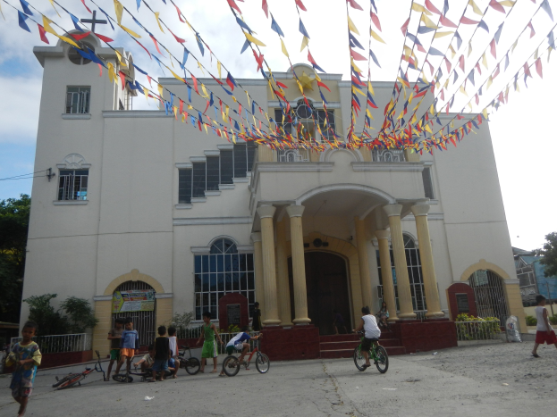 Eine Gruppe von Menschen, die vor einer weißen Kirche mit architectural details, Pflanzen, einem Zaun, einer Fahne, Bäumen und einem bewölkten Himmel Fahrrad fährt, mit bunten Fahnen, die an einem Seil befestigt sind.