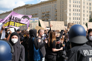 Gruppe maskierter Demonstranten mit Schildern vor einem Gebäude mit zwei Polizisten in Helmen auf der rechten Seite, vor einem bewölkten Himmel mit Bäumen im Hintergrund.