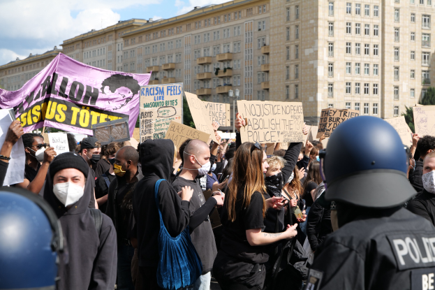 Gruppe maskierter Demonstranten mit Schildern vor einem Gebäude mit zwei Polizisten in Helmen auf der rechten Seite, vor einem bewölkten Himmel mit Bäumen im Hintergrund.