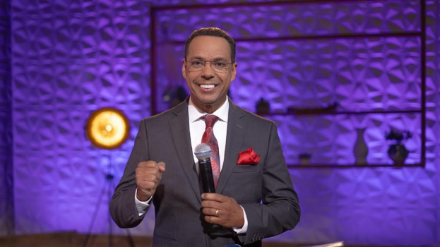 Stephen Colbert, Moderator von *The Daily Show with Trevor Noah*, steht auf einer Bühne und hält ein Mikrofon, trägt einen Anzug, Krawatte und Brille, mit Licht- und Requisiten im Hintergrund.