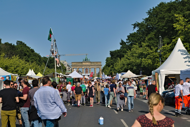 Eine Menschenmenge, die eine Straße entlanggeht, gesäumt von Zelten, Fahrzeugen und Bäumen, mit einem Bogen und einem klaren blauen Himmel im Hintergrund und Masten mit Fahnen auf der linken Seite, wahrscheinlich das Oktoberfest in München, Deutschland.