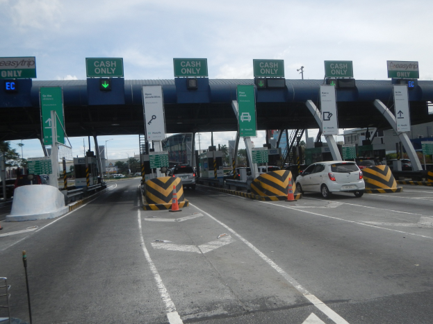 Mautstelle mit darunter fahrenden Fahrzeugen, umgeben von Verkehrskegeln, Absperrpollern, Hinweisschildern, einem Tor, Laternenmasten, Bäumen, Gebäuden und einem bewölkten Himmel.