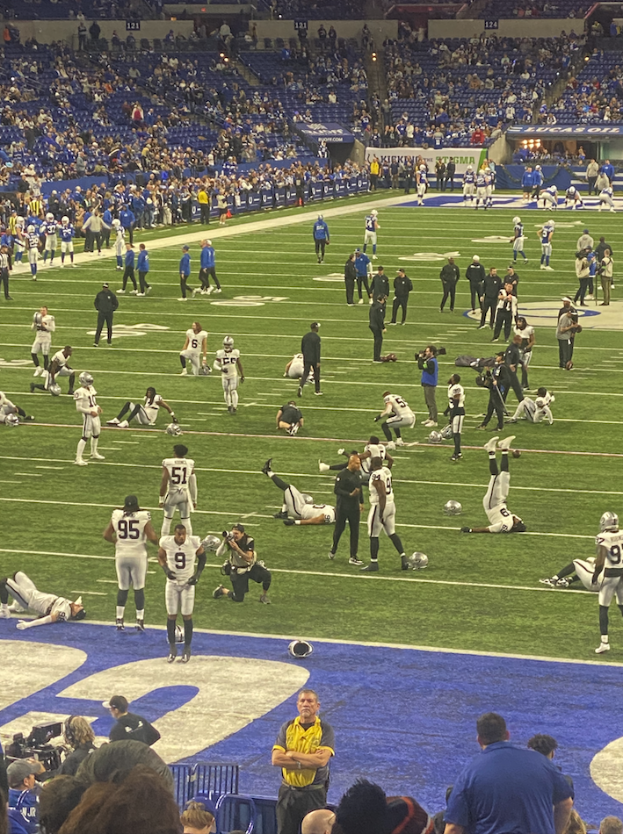 Ein Fußballspiel wird in einem Stadion mit Menschen auf dem Feld und einigen auf Stühlen sitzend im Hintergrund gespielt, während eine Person eine Kamera hält, was auf eine Live-Übertragung hinweist.