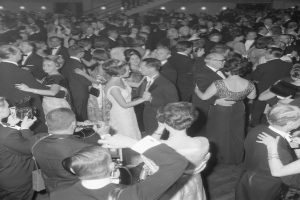 Ein Schwarz-Weiß-Foto von einer Gruppe von Menschen auf einer Tanzfläche, einige halten Kameras, mit Treppen und Lichtern im Hintergrund.