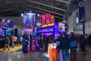 Eine große Gruppe von Menschen steht vor einem Stand auf einer Messe, wobei einige Taschen halten und eine Wand auf der rechten Seite des Bildes zu sehen ist. Der Stand zeigt Tafeln mit der Aufschrift "Indie Game Development Award 2019", Bildschirme, Lampen und andere Gegenstände.