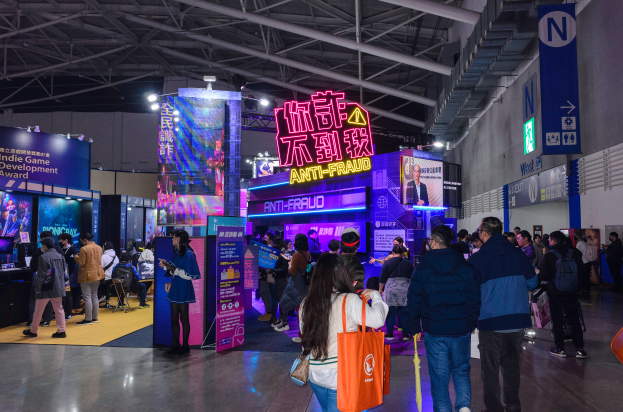 Eine große Gruppe von Menschen steht vor einem Stand auf einer Messe, wobei einige Taschen halten und eine Wand auf der rechten Seite des Bildes zu sehen ist. Der Stand zeigt Tafeln mit der Aufschrift "Indie Game Development Award 2019", Bildschirme, Lampen und andere Gegenstände.