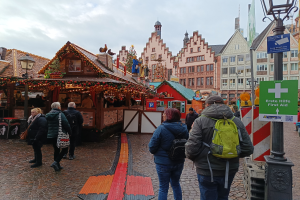 Eine Gruppe von Menschen geht eine Kopfsteinpflasterstraße entlang an einem Weihnachtsmarkt in Nürnberg, Deutschland, vorbei mit Laternenmasten, Texttafeln und Gebäuden mit Fenstern im Hintergrund unter einem bewölkten Himmel.