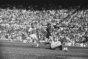 Schwarzes und weißes Foto von Männern, die auf einem Feld Fußball spielen, mit Zuschauern im Hintergrund und einer 'Bundesliga 1978-79 Ajax 1-1 Hannover 96'-Tafel.