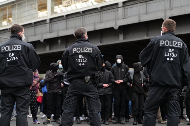 Eine Gruppe von Polizisten in Uniform steht vor einer Menge von Menschen, die ebenfalls schwarze Uniformen und Masken tragen, mit einer Brücke und einem Gebäude im Hintergrund, während einer Demonstration in einer Stadt.