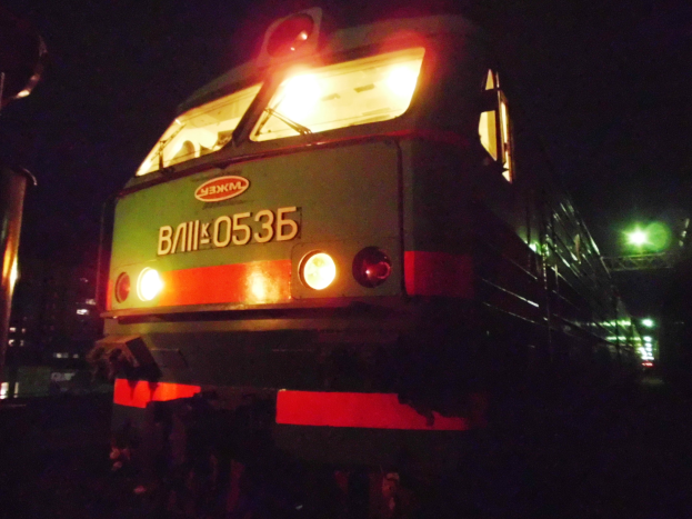 Ein Zug fährt nachts auf beleuchteten Schienen, mit Laternen auf der linken Seite und einem dunklen Himmel im Hintergrund.