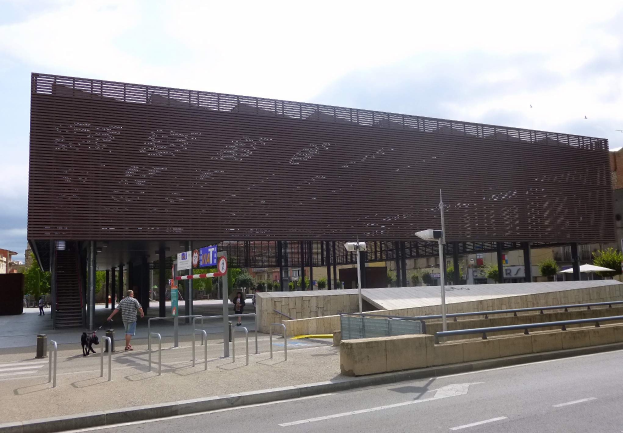 Großes modernes Gebäude mit zahlreichen Fenstern, das als neuer Eingang zur Stadt dient, umgeben von Straßeninfrastruktur, Fahrzeugen, Fußgängern und Bäumen unter einem bewölkten Himmel.